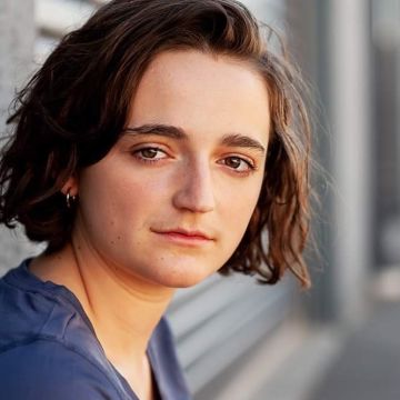 Woman with short brown hair, leaning against a concrete wall and looking to camera