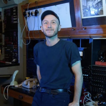 A man with light hair is wearing a hat and a blue shirt. He is leaning against a desk while smiling and looking to camera