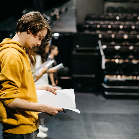 A teenage drama student, wearing a yellow sweater holds a drama script, while standing in The Roundhouse theatre, surrounded by other young drama students 