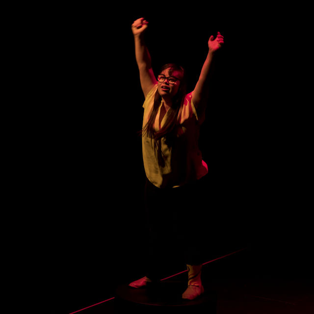 Full body image of woman standing on a dark stage, looking up with her hands in the air