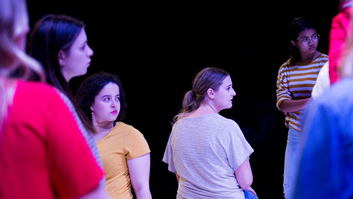 A photo captures school drama students, in the middle of a production at La Boite Theatre. There are a number of them on stage, set against a black background and lit with a blue light. 