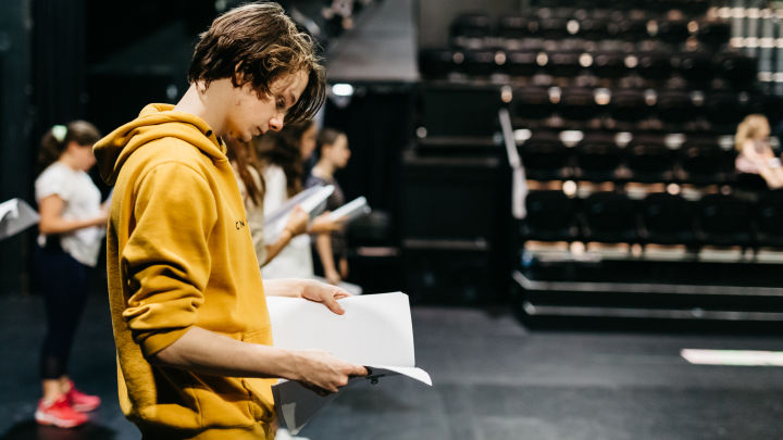 A teenage drama student, wearing a yellow sweater holds a drama script, while standing in The Roundhouse theatre, surrounded by other young drama students 