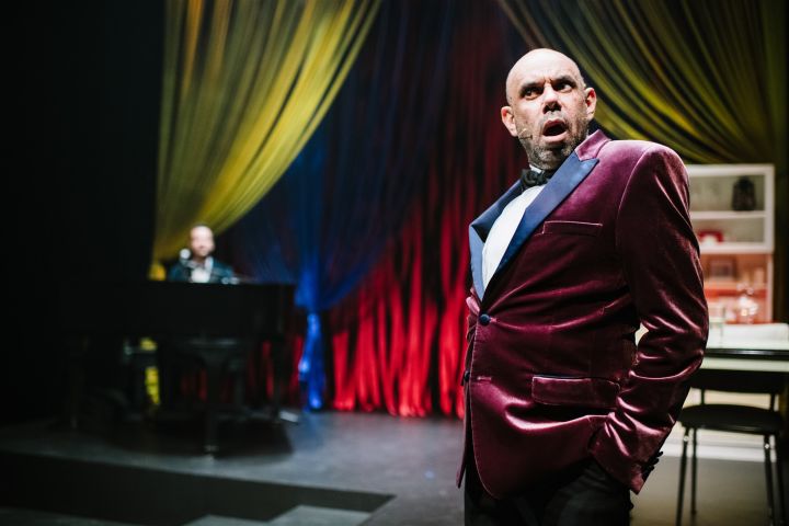 Image of a man standing on stage with a stunned expression. He is wearing a tuxedo made out a red velvet material. There is a pianist in the background accompanying him for the theatre show. This show has been recorded to deliver online theatre for studen