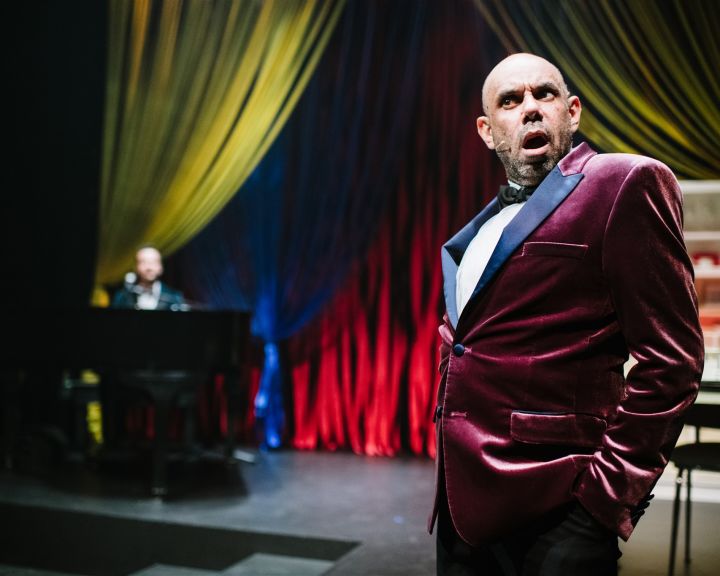 Image of a man standing on stage with a stunned expression. He is wearing a tuxedo made out a red velvet material. There is a pianist in the background accompanying him for the theatre show. This show has been recorded to deliver online theatre for studen